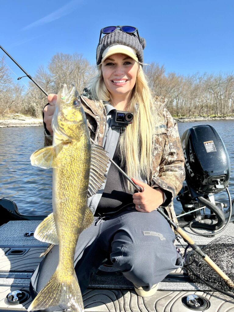 A female angler kneels on a boat, holding a large walleye she has caught. She is smiling and looking at the camera. She is wearing a camouflage jacket, dark pants, a knit beanie, and sunglasses. A GoPro camera is visible on her chest. The boat is on a body of water with trees on the distant shore under a clear blue sky. A fishing rod is visible in the foreground and a boat motor in the background.