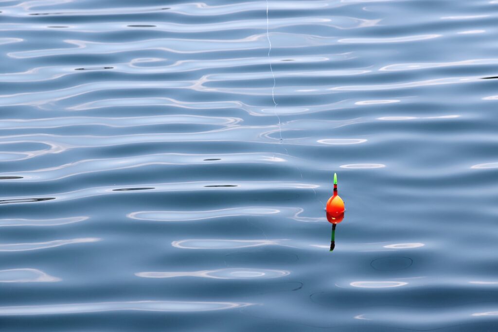 An orange and green fishing bobber floats on the rippled surface of blue-grey water, with a fishing line extending upwards from it.