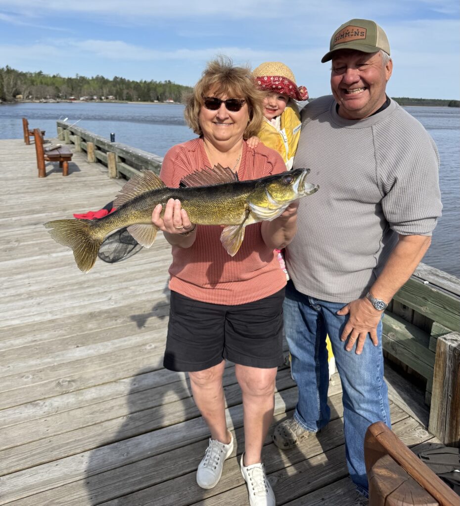 An older woman, an older man, and a young child stand on a wooden dock, proudly displaying a large walleye fish. The woman holds the fish horizontally, smiling. The man stands next to her, also smiling, wearing a baseball cap. The child is partially visible behind the woman, wearing a hat. In the background, a lake or body of water stretches out, with a distant shoreline featuring houses and trees under a partly cloudy sky. This alt text is optimized for SEO with keywords like "walleye fishing," "family fishing," "fishing dock," and "trophy fish."
